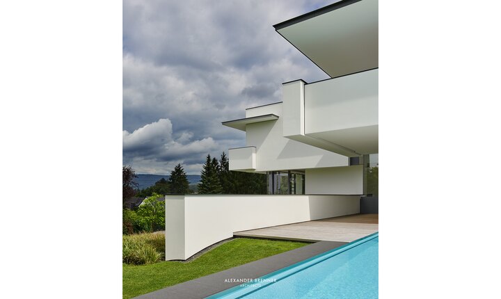 Die hell verputzte Betonfassade im Kontrast zum Grün der angrenzenden Natur. Foto: © Alexander Brenner Architects